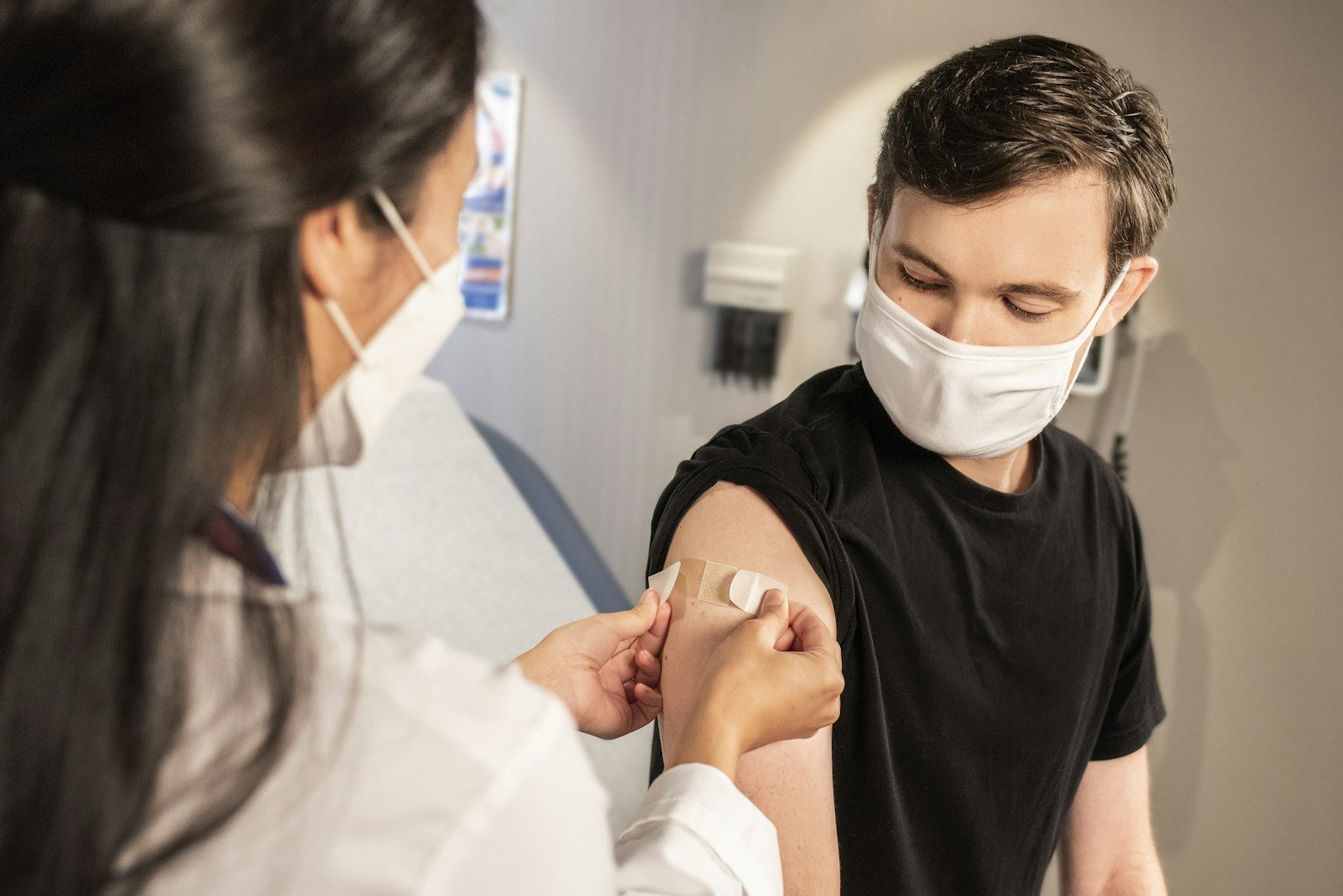 man in a black t-shirt receiving a vaccination from a doctor in white cat in his left arm. Both are people are wearing a face mask.
